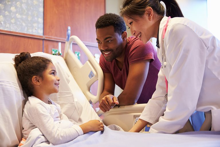 Young girl in hospital bed with father next to her and a doctor speaking with her.
