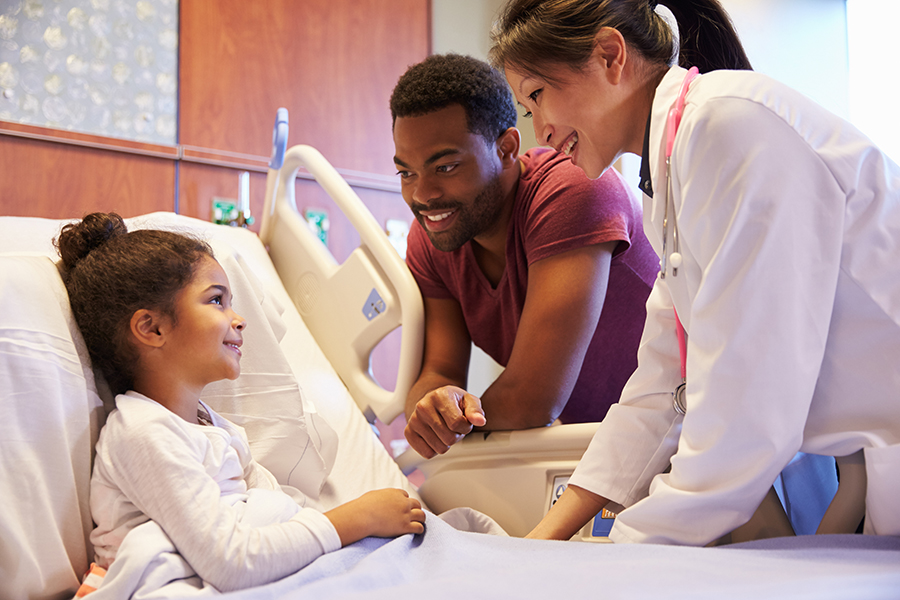 Young girl in hospital bed with father next to her and a doctor speaking with her.