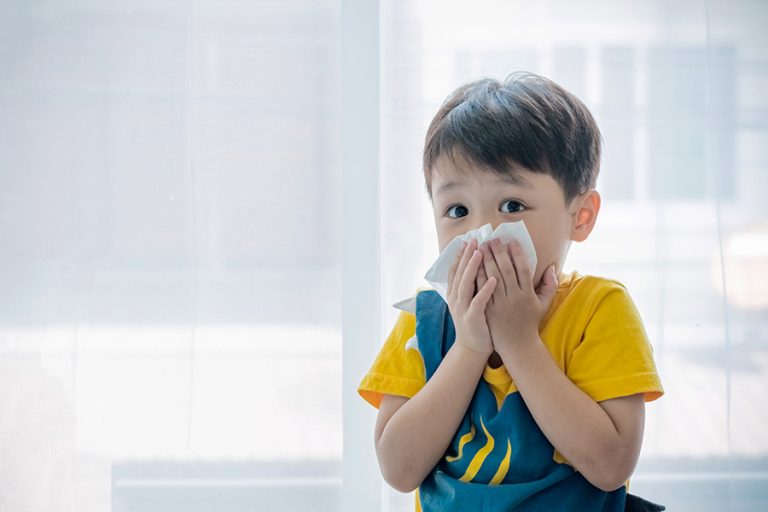 Young boy mid-sneeze holding a tissue to his nose.